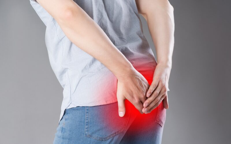 Man suffering from hemorrhoids, anal pain on gray background, painful area highlighted in red